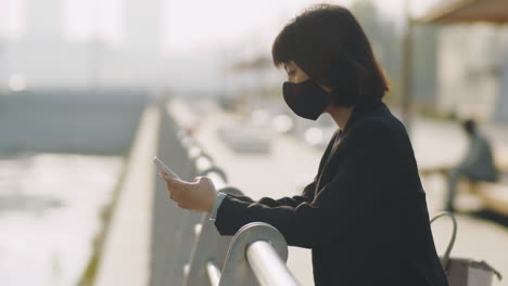 Asian-Businesswoman-in-Mask-Using-Phone-on-Urban-Embankment